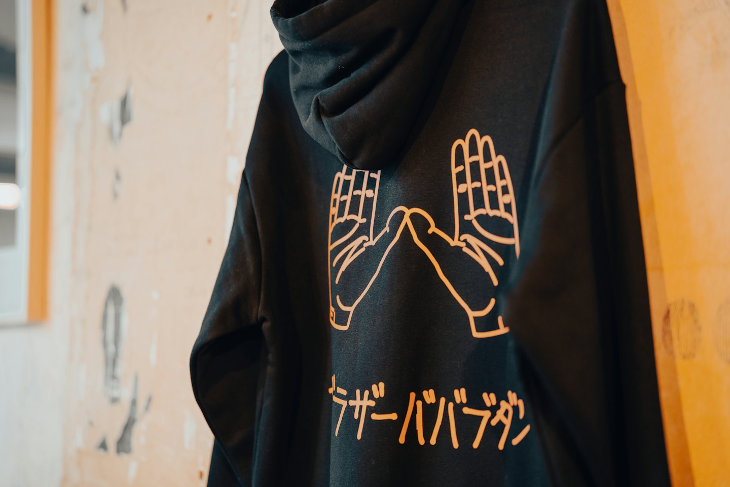 Schwarzer Hoodie mit orangefarbenem Handdesign hängt an einer Wand.